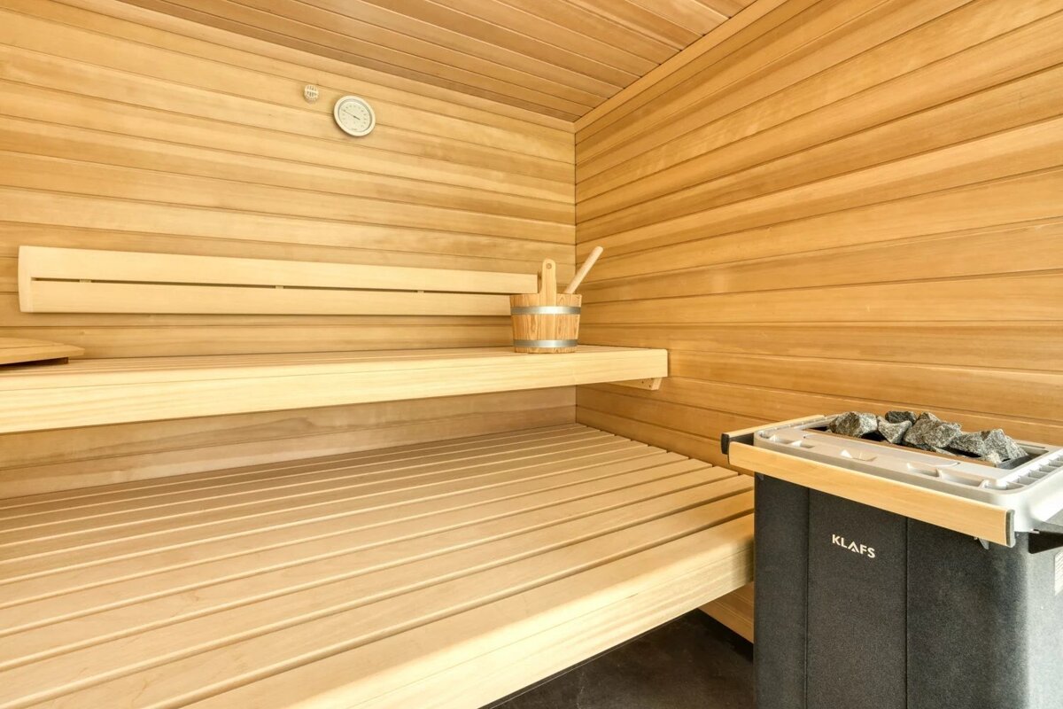 A clean wooden sauna featuring tiered benches, a wooden bucket with a ladle, a wall thermometer, and a Klafs stone heater.