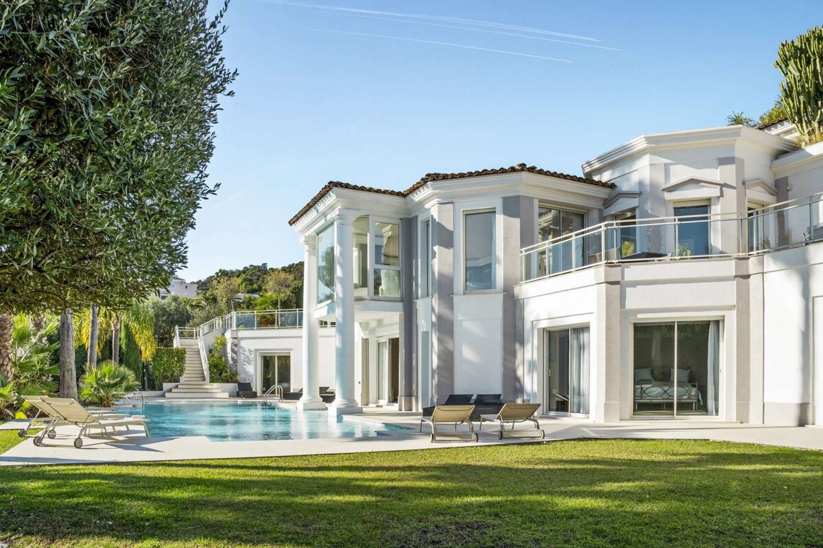 Luxurious white villa with a blue swimming pool, green lawn, and olive trees under a sunny sky. Modern architecture with balconies.