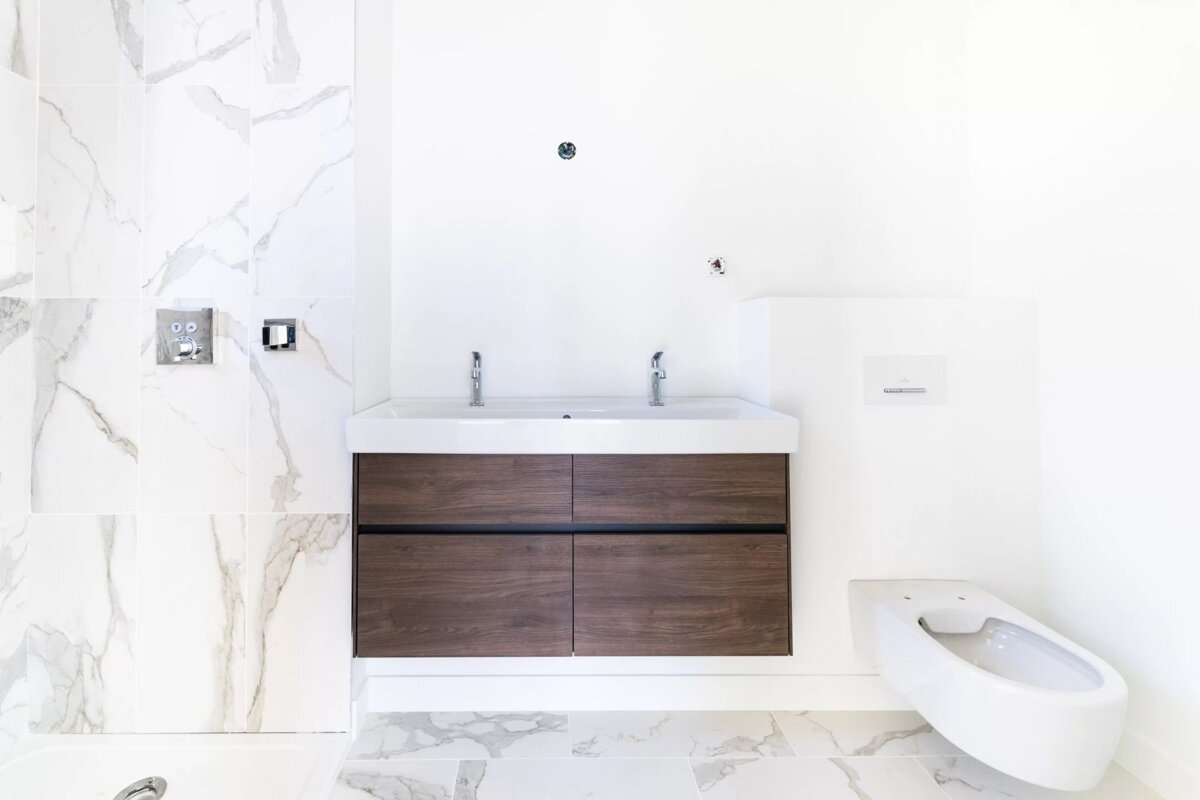 A modern, unfinished white bathroom featuring a marble tiled floor, a wooden vanity with two faucets, and a toilet bowl.