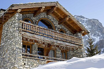 A rustic stone and timber chalet nestled in deep snow, featuring ornate wooden balconies and arched windows, with a snow-capped mountain in the background.