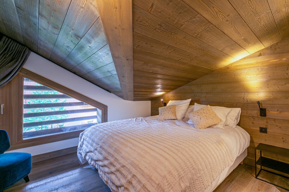 Cozy bedroom with slanted wooden walls & ceiling, a large bed, and an angled window revealing trees outside. Rustic-modern feel.