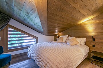 Cozy bedroom with slanted wooden walls & ceiling, a large bed, and an angled window revealing trees outside. Rustic-modern feel.