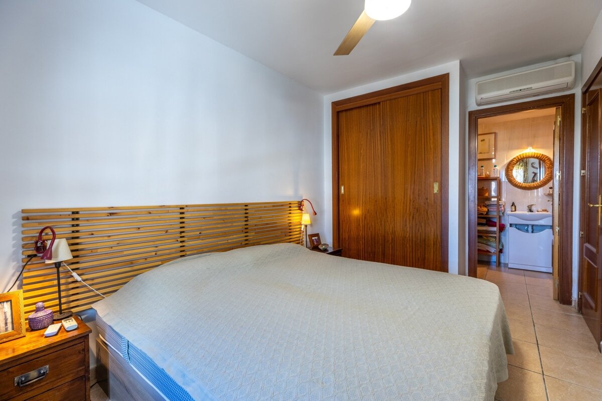 A bedroom with a large bed, wooden headboard, built-in wardrobe, and an open doorway to a bathroom. Features include a ceiling fan and AC unit.