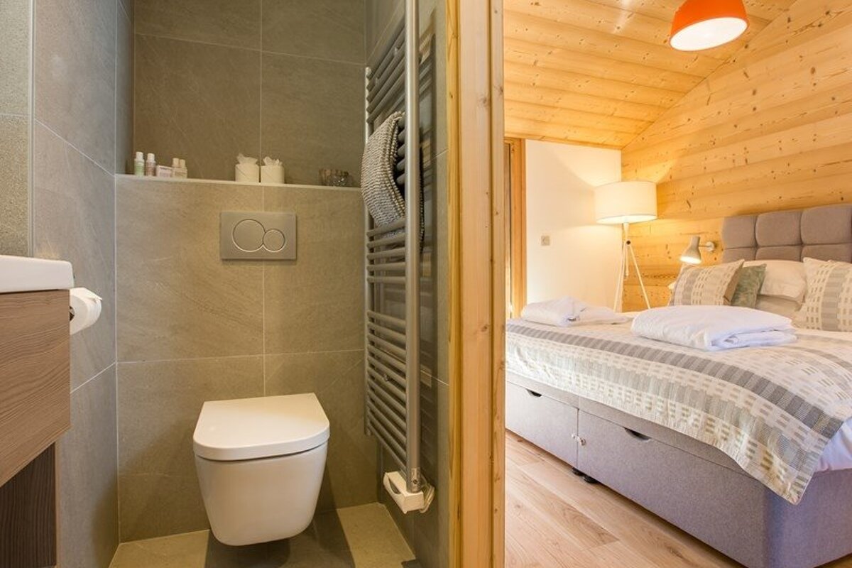 A modern tiled bathroom with a wall-mounted toilet, looking into a cozy cabin bedroom with light wood walls and a bed.