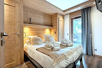 Cozy, light wood paneled bedroom featuring a bed with fluffy pillows and bathrobes, plus a balcony door and warm accent lighting.