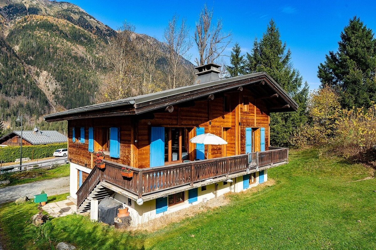 A charming wooden chalet with striking blue shutters and a balcony, surrounded by green grass, with mountains and pine trees in the background.