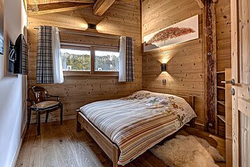 A cozy, rustic wood-paneled bedroom with a bed, window, wall-mounted TV, and furry rug. Exposed beams enhance its warm, cabin-like atmosphere.
