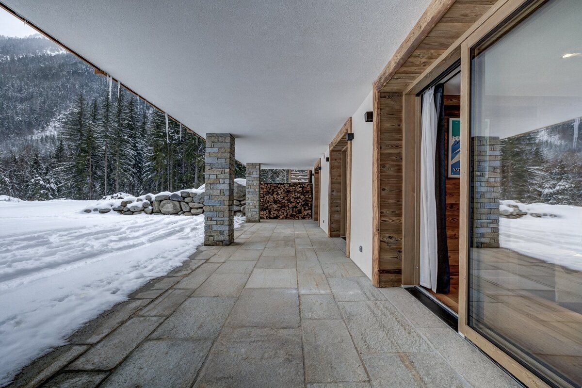 A modern chalet's covered stone patio extends into a snowy landscape with a forested mountain backdrop. Icicles hang, and firewood is neatly stacked.