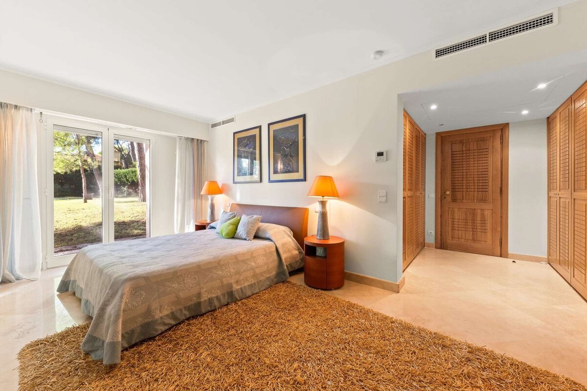 A bright bedroom with a bed, shaggy orange rug, two lamps, and glass doors leading to a garden. Wooden built-in closets and a door are visible.