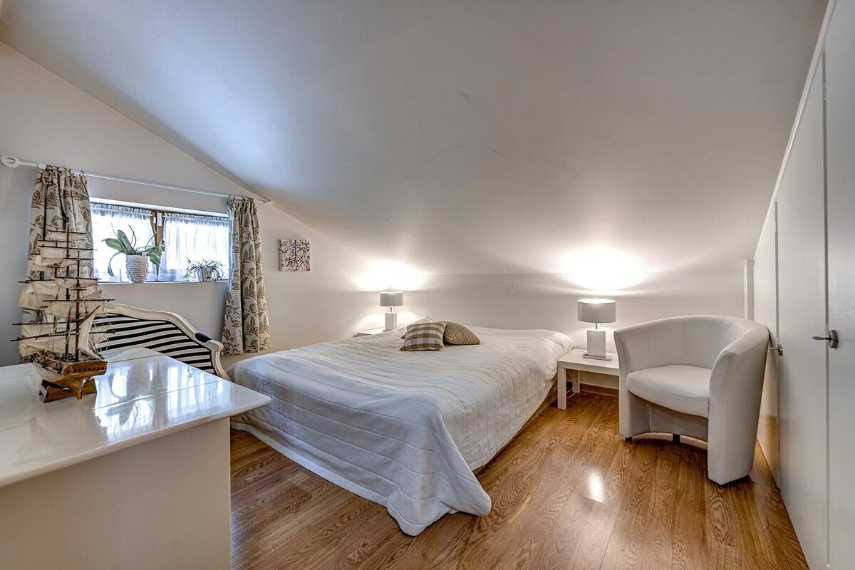 Bright attic bedroom with a white bed, sloped ceiling, wooden floor, window, and a ship model on a table.