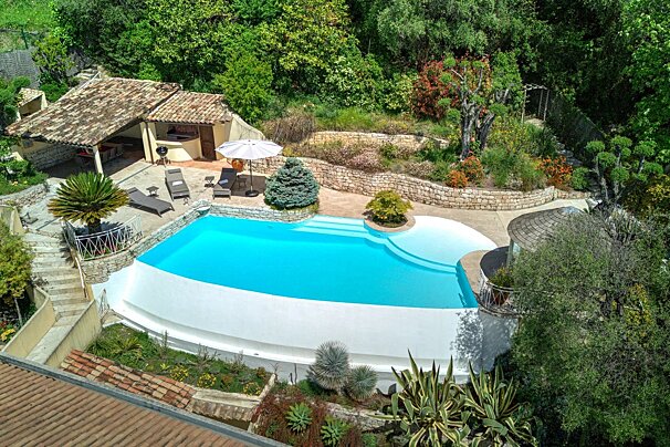 Aerial view of a luxury backyard featuring a bright blue swimming pool, sun loungers, an outdoor patio, and lush green gardens.