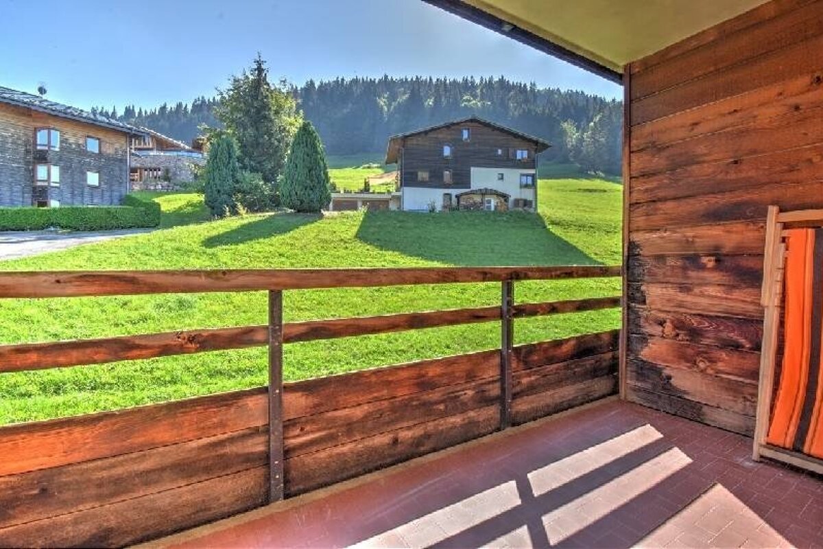 View from a rustic wooden balcony overlooking green alpine hills, chalets, and pine trees.