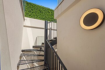 An outdoor black metal spiral staircase between white walls, featuring a glowing circular light fixture and a green hedge at the top.