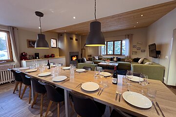 Warm chalet interior: long dining table set for guests, green L-shaped couch, and a cozy fireplace, with snow visible outside.
