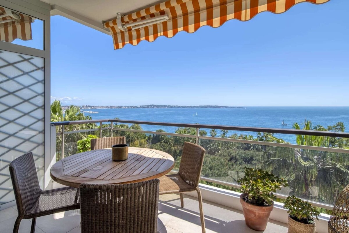 Sunny balcony with a wooden table, chairs, and a striped orange awning, offering a stunning panoramic view of the blue ocean and distant coastline.