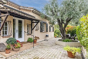 A cream-colored house with a tiled roof, white door, and shutters, beside a paved patio with a large olive tree and outdoor seating.