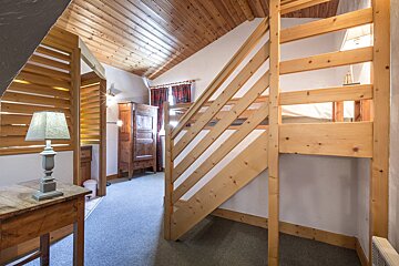 A cozy, rustic room features a wooden slatted ceiling, a small table with a lamp, an antique wardrobe, and light wood stairs/bunk beds.