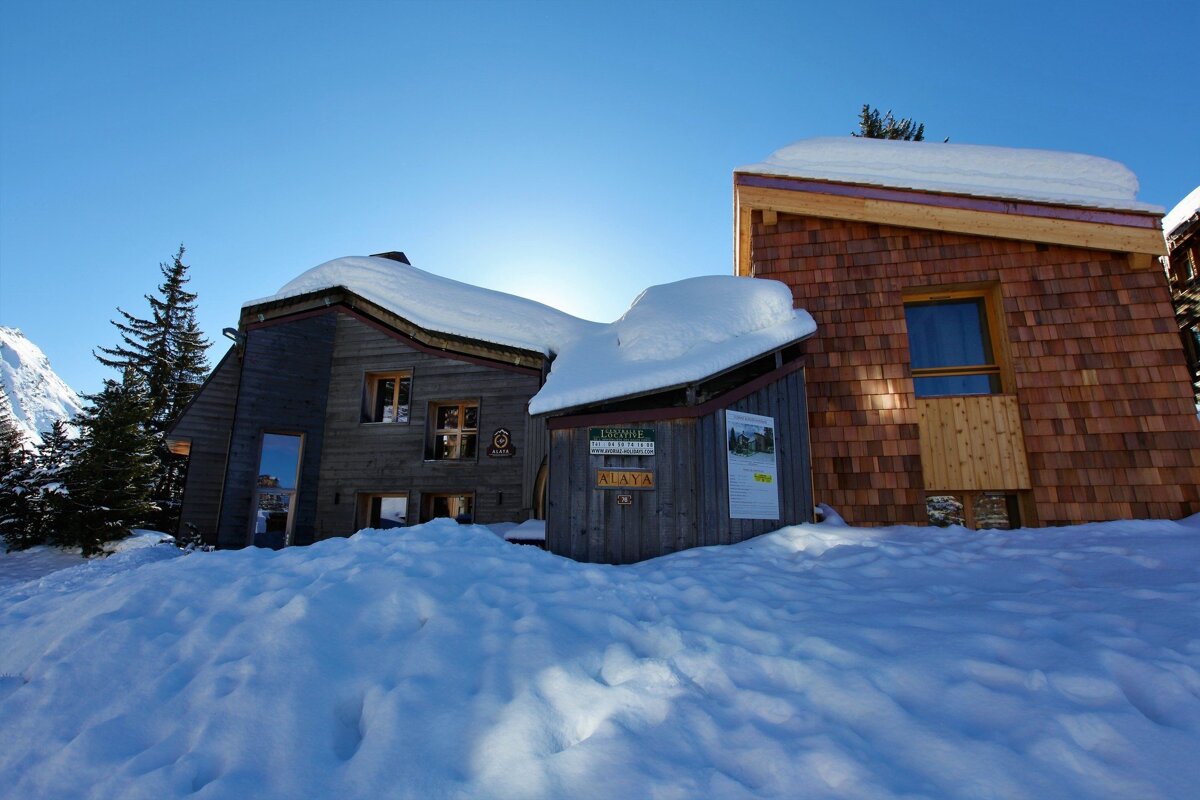 Two snow-covered wooden chalets with a bright blue winter sky overhead. Deep snow covers the ground, and distant mountains and trees are visible.