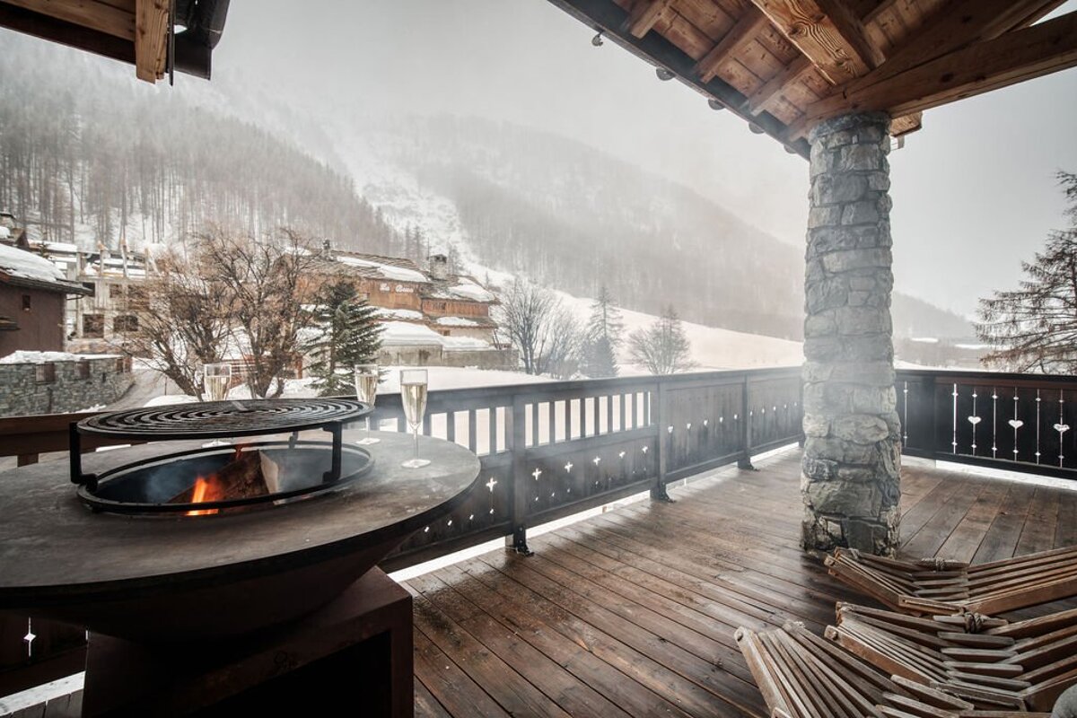A cozy winter chalet balcony with a lit fire pit, champagne flutes, and snowy mountain village views under a misty sky.