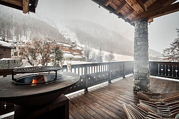 A cozy winter chalet balcony with a lit fire pit, champagne flutes, and snowy mountain village views under a misty sky.