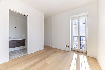 Bright, empty room with white walls, light wood floor, balcony doors, and a doorway revealing a modern bathroom vanity.