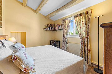A cozy bedroom features yellow walls, exposed wooden beams, a large bed with floral pillows, and a window with patterned curtains. Bookshelves hold many books.