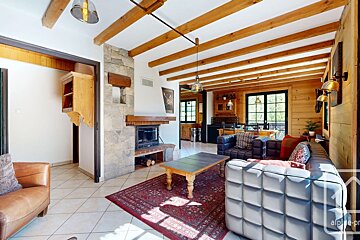 Rustic living room with exposed wood beams, stone fireplace, tiled floor, red rug, and leather sofas, leading to a bright kitchen/dining area.