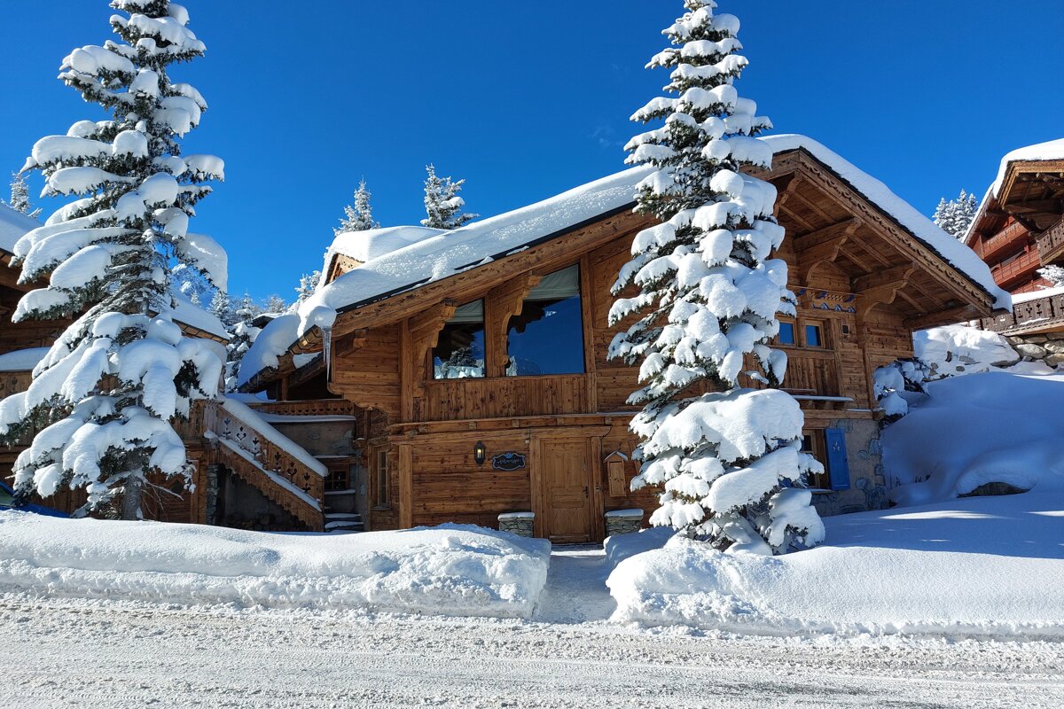 A cozy wooden chalet and snow-laden pine trees stand under a bright blue sky on a clear, sunny winter day.