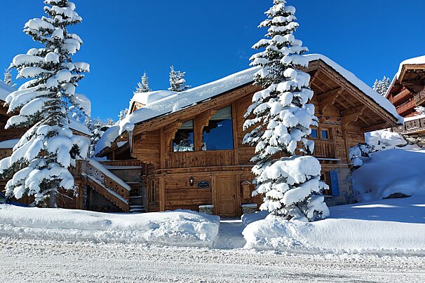 A cozy wooden chalet and tall pine trees blanketed in fresh snow against a bright blue winter sky, depicting a picturesque winter scene.