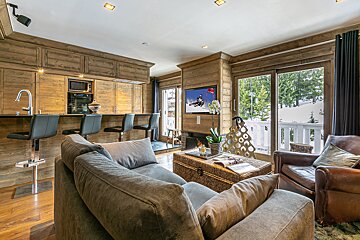 A cozy, modern chalet-style living room and kitchen area with wooden walls, comfortable seating, a TV showing skiing, and a snowy mountain view.