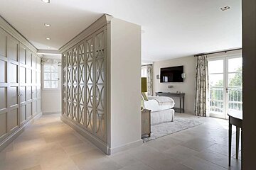A modern, bright bedroom with a walk-in closet/dressing area. It features light-toned decor, large windows, and a wall-mounted TV.