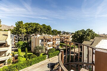 View from a spiral balcony over residential buildings with tiled roofs, lush trees, and distant mountains under a clear blue sky.