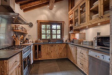 Rustic kitchen featuring natural wood cabinetry, exposed ceiling beams, a large window overlooking trees, and stainless steel appliances.