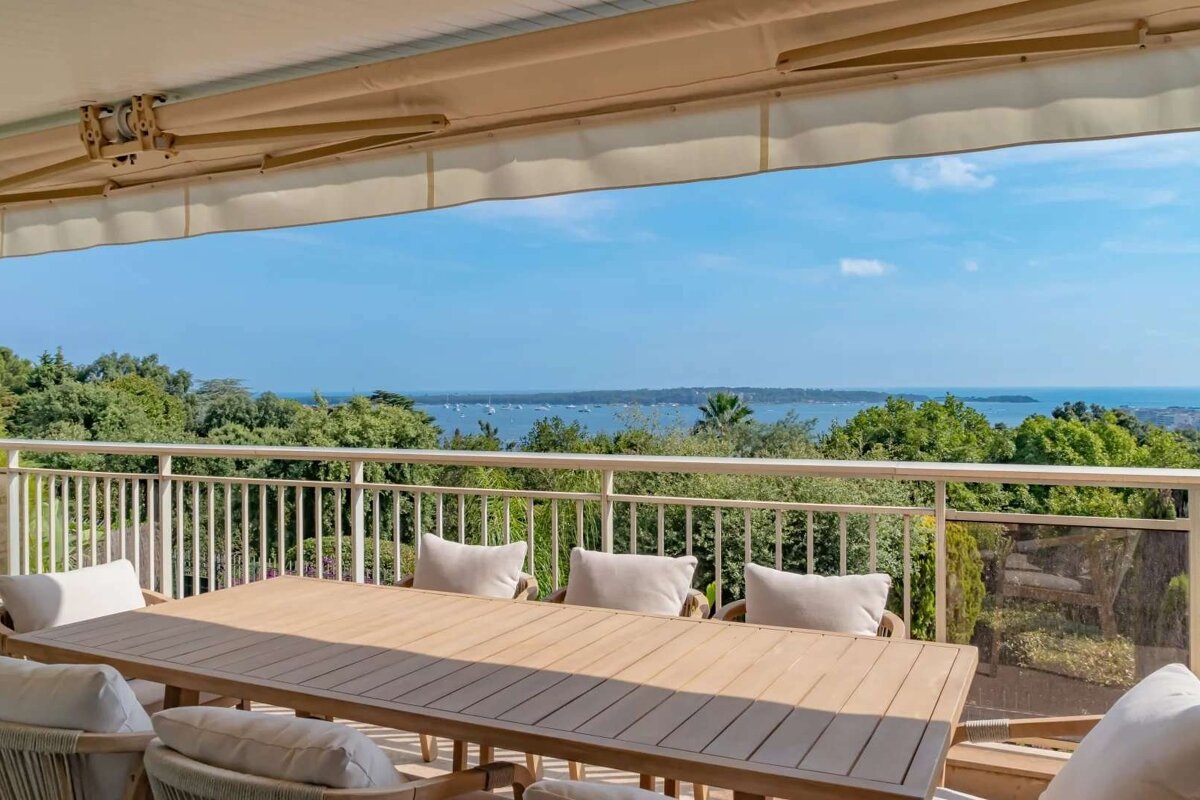 Sunny balcony with an outdoor dining set, offering a stunning panoramic view of a blue sea with boats, green trees, and a clear sky.