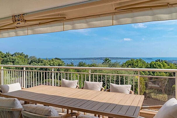 Sunny terrace with a wooden dining set, offering panoramic views of a blue sea dotted with boats and lush green trees below.