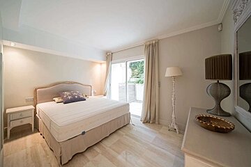 A bright, neutral-toned bedroom features a large bed, nightstand, dresser with lamps, and sliding glass doors opening to a patio.
