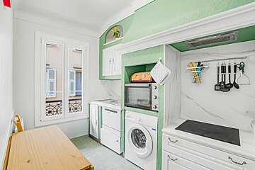 Compact kitchen featuring light green and white cabinets, a washing machine, oven, a small wooden table, and a window.