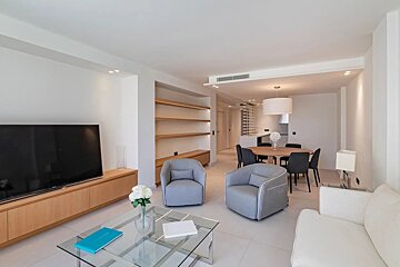 Bright, modern apartment with a minimalist living room featuring grey armchairs, a large TV, and an open dining area in the background.