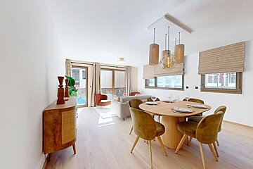 A modern open-plan living/dining room featuring a round wood table with olive velvet chairs, a light sofa, stylish chandelier, and large windows on light wood floors.