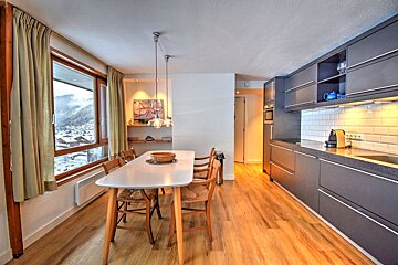 Modern open-plan kitchen and dining area with a long table, sleek dark cabinets, and a large window showcasing a snowy mountain view.