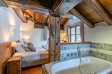 Rustic bedroom with exposed wooden beams, a comfy bed, a large window, and a unique built-in bathtub with tiled surround.