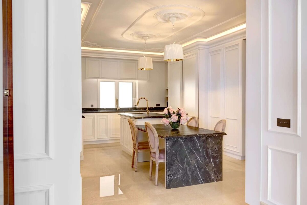 An elegant white kitchen with a black marble island, ornate ceiling, and pink floral centerpiece, viewed through an open doorway.