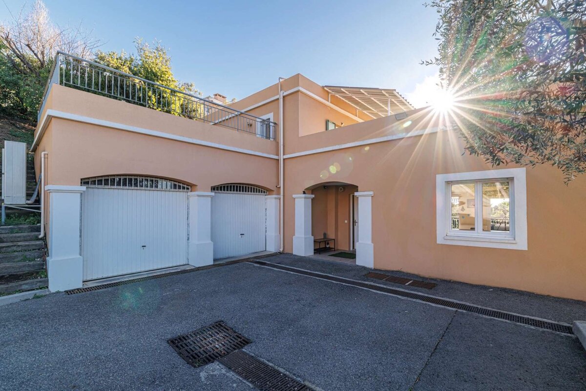A peach-colored house with two white garage doors and an arched entry under a bright blue sky with strong sun flare over a paved driveway.