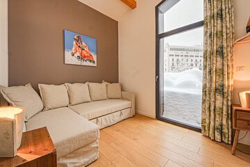 A cozy room features a beige L-shaped sofa, a unique painting of a skiing dog, warm wooden floors, and a large window overlooking a snowy landscape.