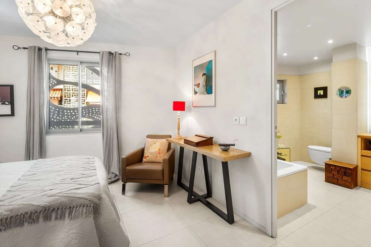A modern bedroom with a bed, armchair, and console table. An open doorway reveals an adjoining bathroom with a toilet and tub. Unique window decor.