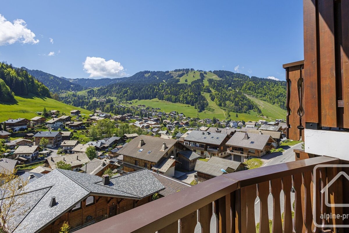 View from a balcony overlooking a scenic alpine village nestled in a lush green valley with forested mountains and ski slopes under a clear blue sky.