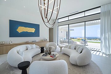 Bright, modern living room with white curved sofas, abstract artwork, and a unique chandelier. Floor-to-ceiling windows reveal a vast outdoor landscape and ocean.
