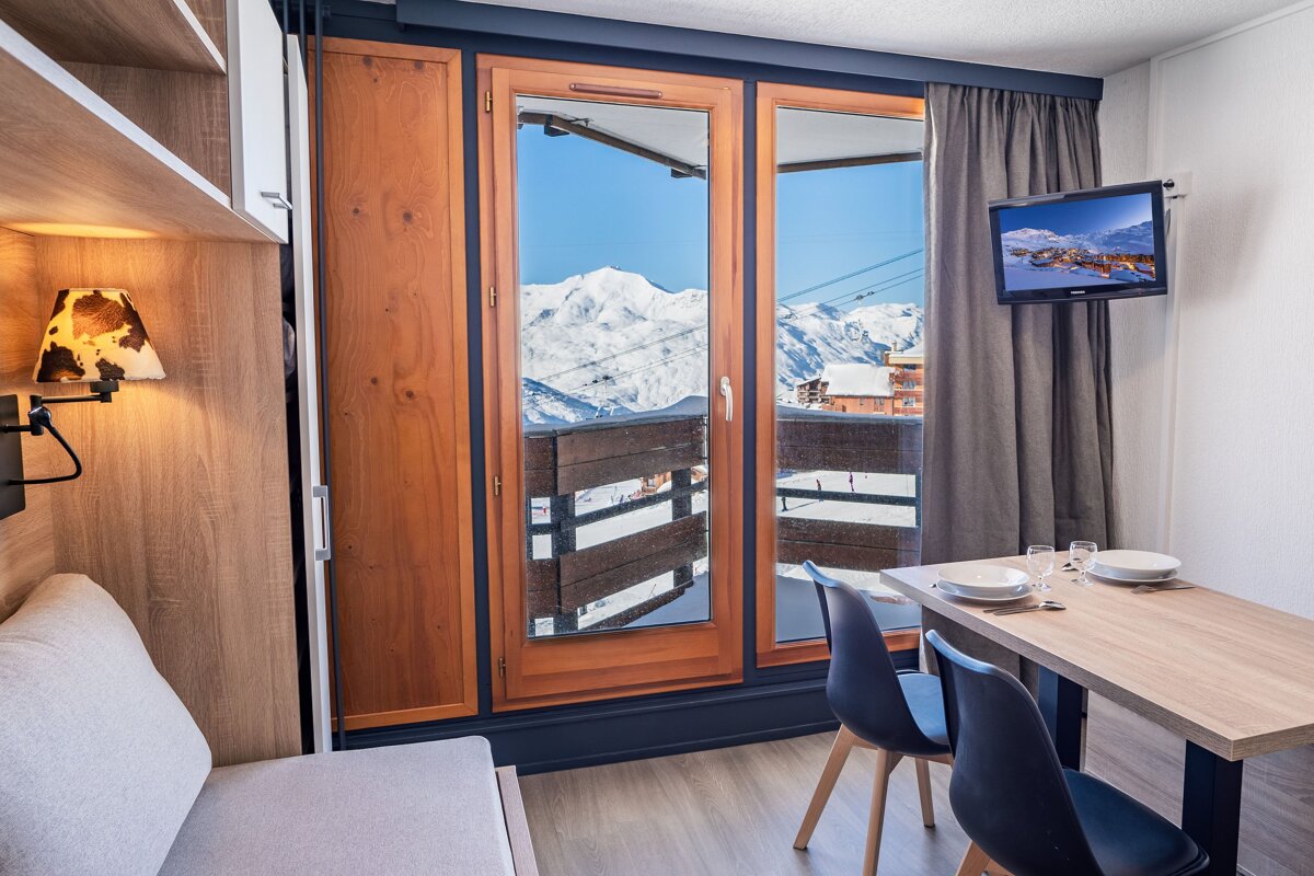 A cozy ski apartment room featuring a dining table, couch, wall-mounted TV, and a stunning balcony view of snow-covered mountains and ski slopes.