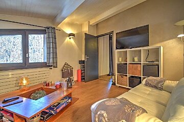 Warm living room featuring a white leather sofa, wooden coffee table, window, and a wall-mounted TV above a white cube shelf.