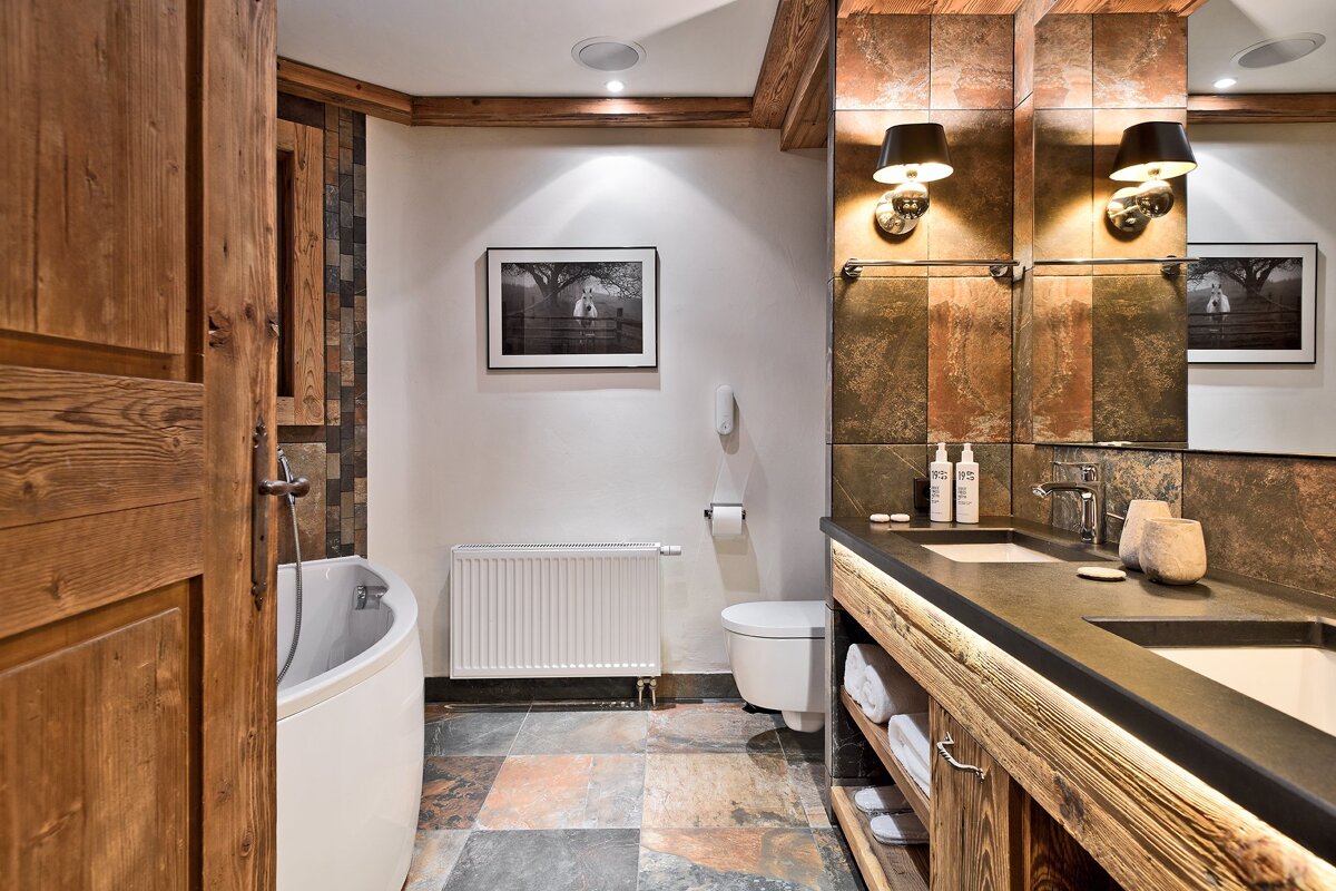A rustic bathroom features a white tub, toilet, and double vanity with wood and multi-colored stone tile accents. A framed black and white horse photo hangs on the wall.